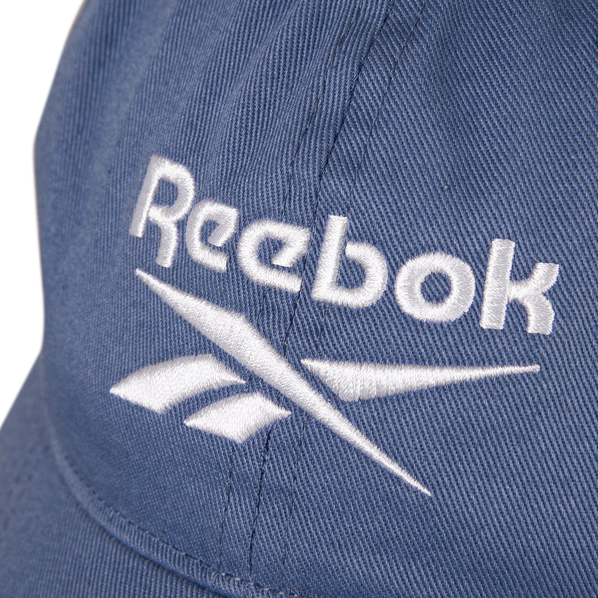 Close-up of the Reebok Logo Cap, made from cotton twill with a visible white embroidered Reebok logo and text. The cap features fabric texture details and an adjustable buckle.