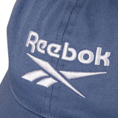 Close-up of the Reebok Logo Cap, made from cotton twill with a visible white embroidered Reebok logo and text. The cap features fabric texture details and an adjustable buckle.