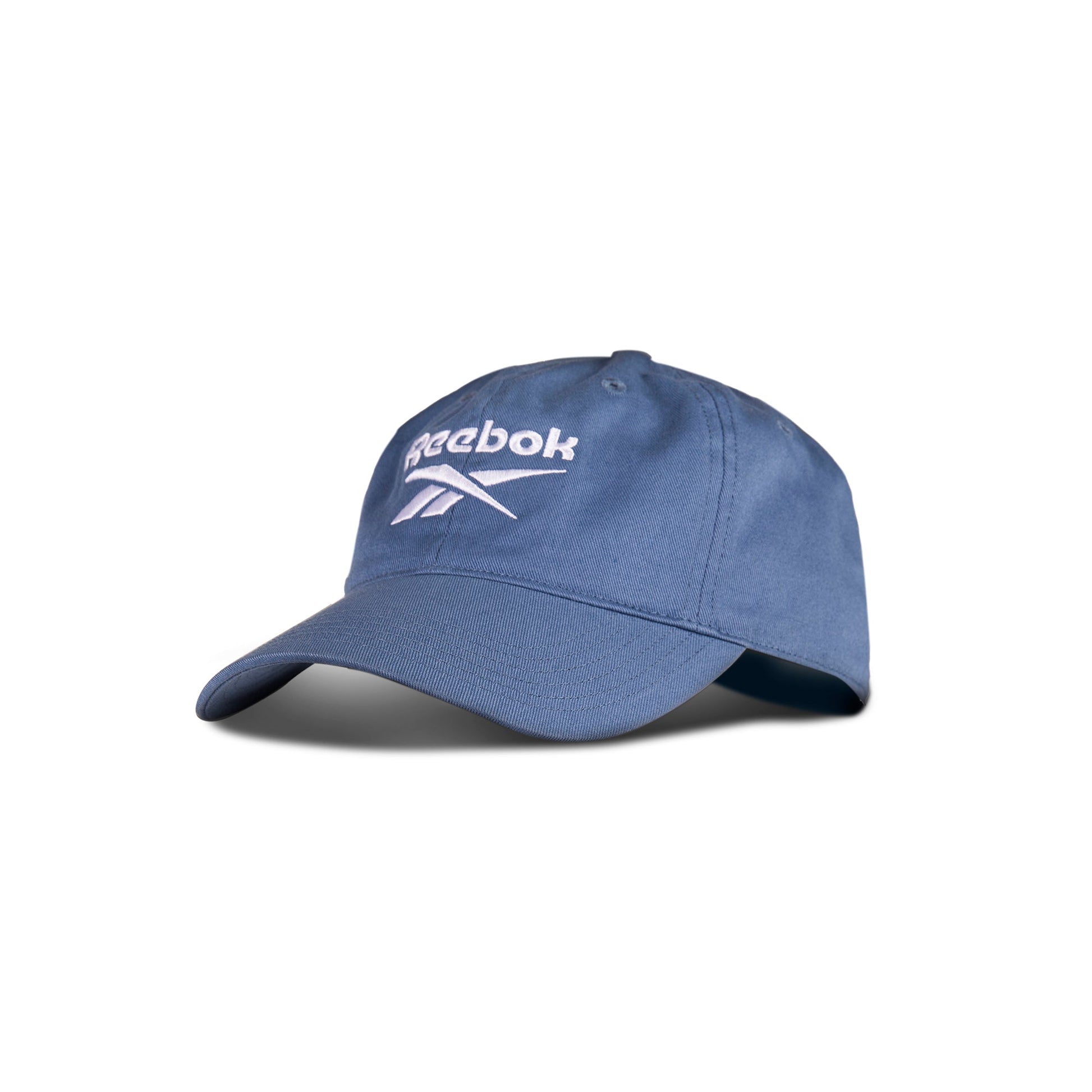 The Reebok Logo Cap is a blue cotton twill cap with a curved brim, white Reebok logo and name on the front, and an adjustable buckle for comfort. Shown against a plain white background.