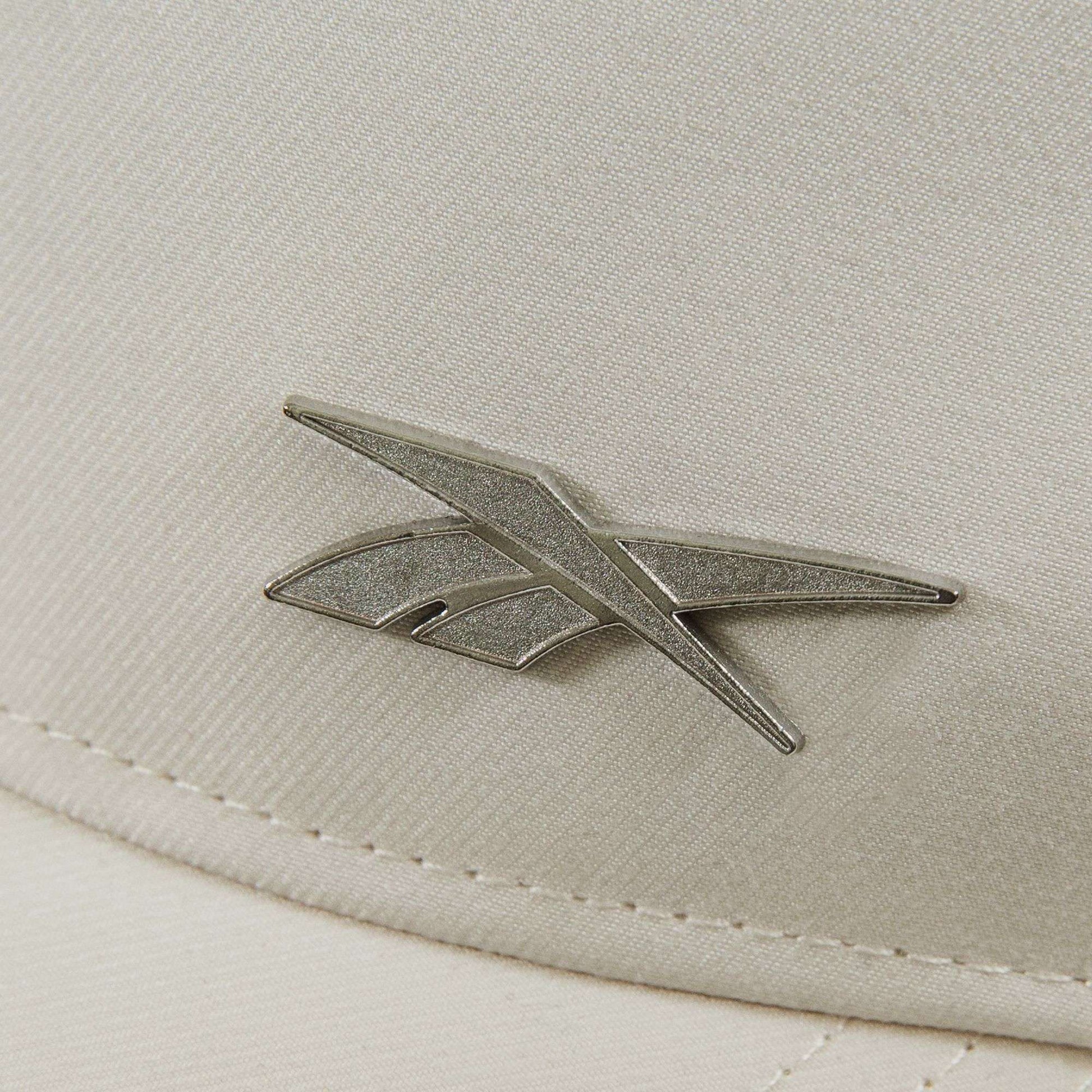 Close-up of the Reebok Badge Cap shows its beige fabric surface with a metallic angular Reebok logo and visible stitching along the edge.