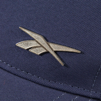 A close-up of a metallic Reebok logo pin on the Badge Cap, featuring dark blue fabric with detailed stitching and a shiny logo.