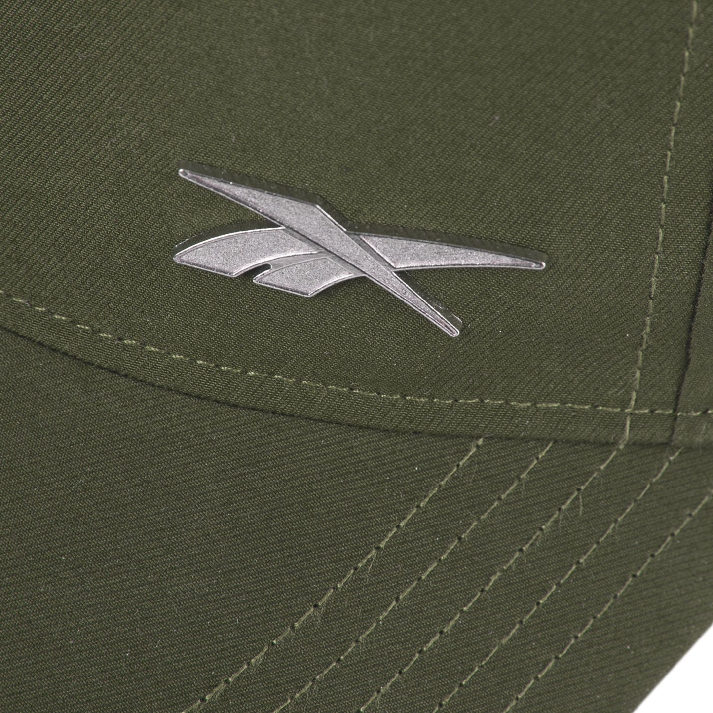 Close-up of the silver Reebok logo on olive green fabric of the Reebok Badge Cap, featuring visible stitching and intersecting seams.