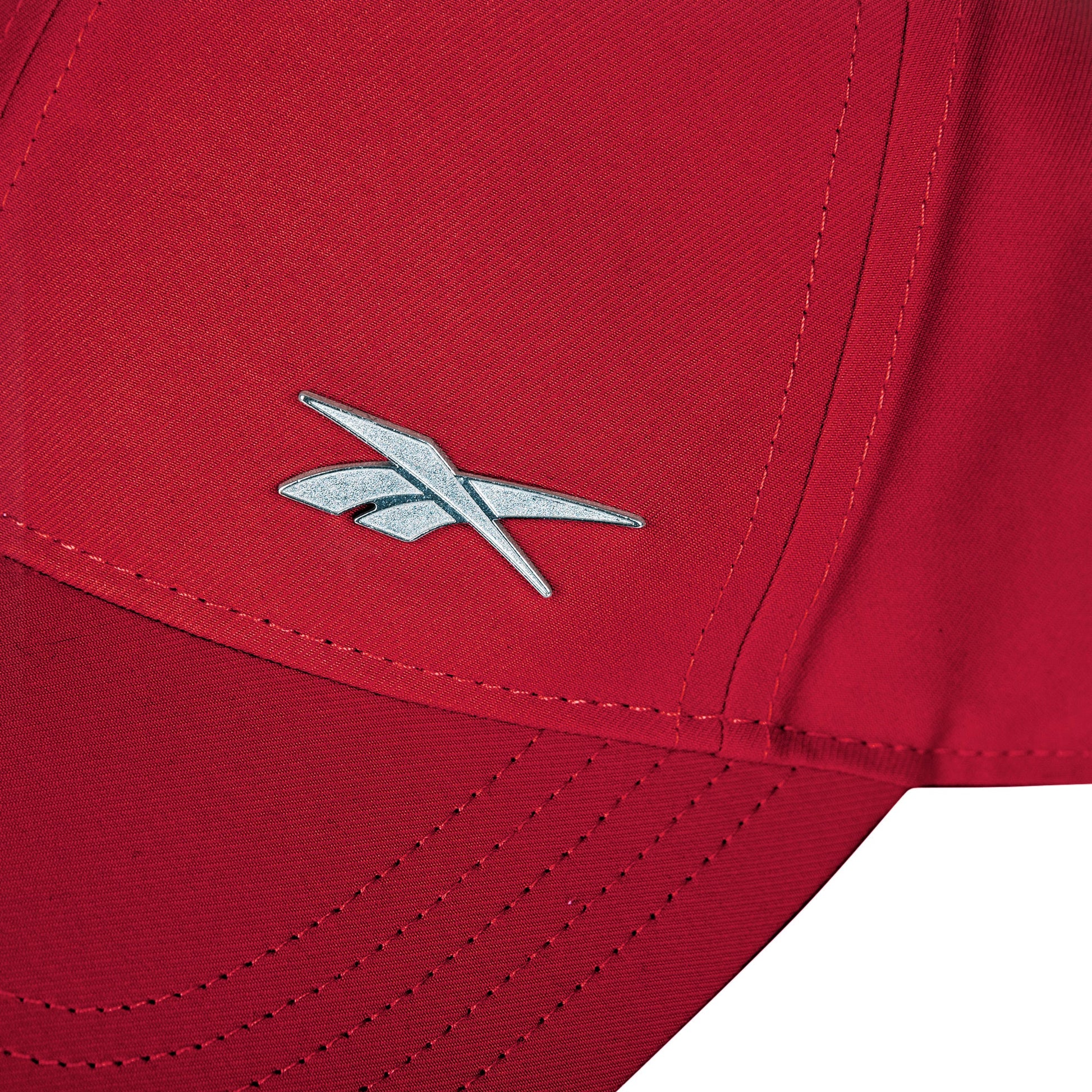 Close-up of the Reebok Badge Cap in red, showcasing a silver logo on the front panel, detailed seams, and an adjustable buckle for a customized fit.