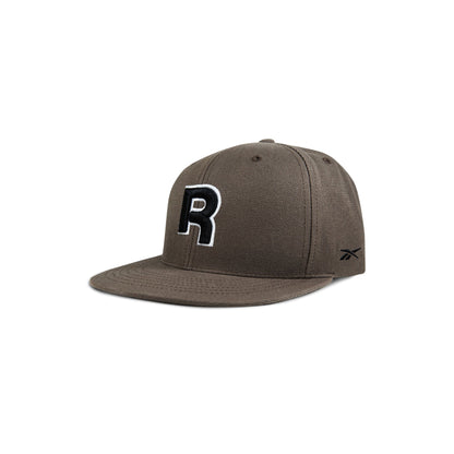 The Reebok Identity Drop R Cap features a flat brim, large black and white R on the front, small side logo, and a snapback closure. Shown in brown on a white background.
