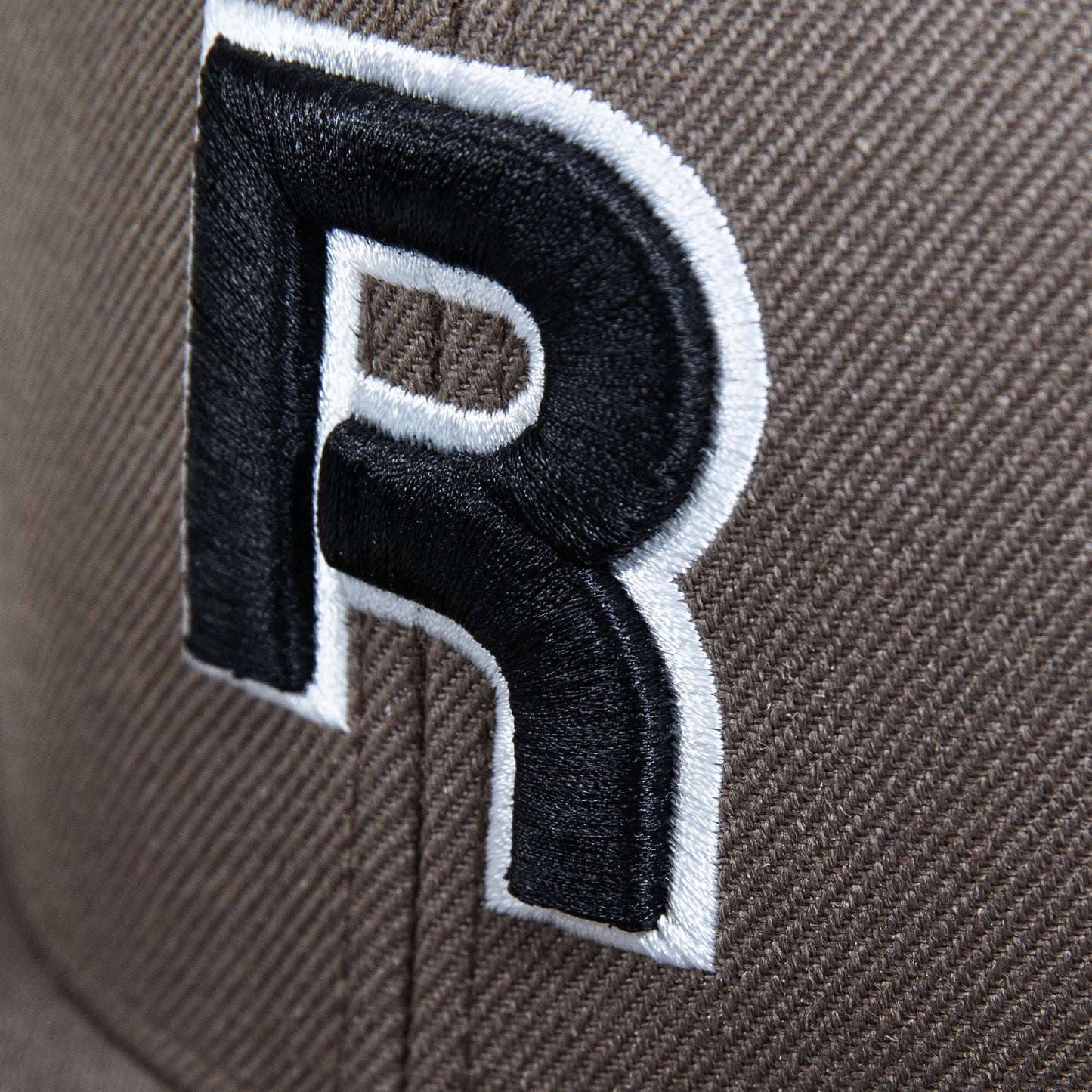 Close-up of Reebok's Identity Drop R Cap in brown, showcasing a large embroidered black "R" outlined in white, with visible stitching, texture, and a flat brim.