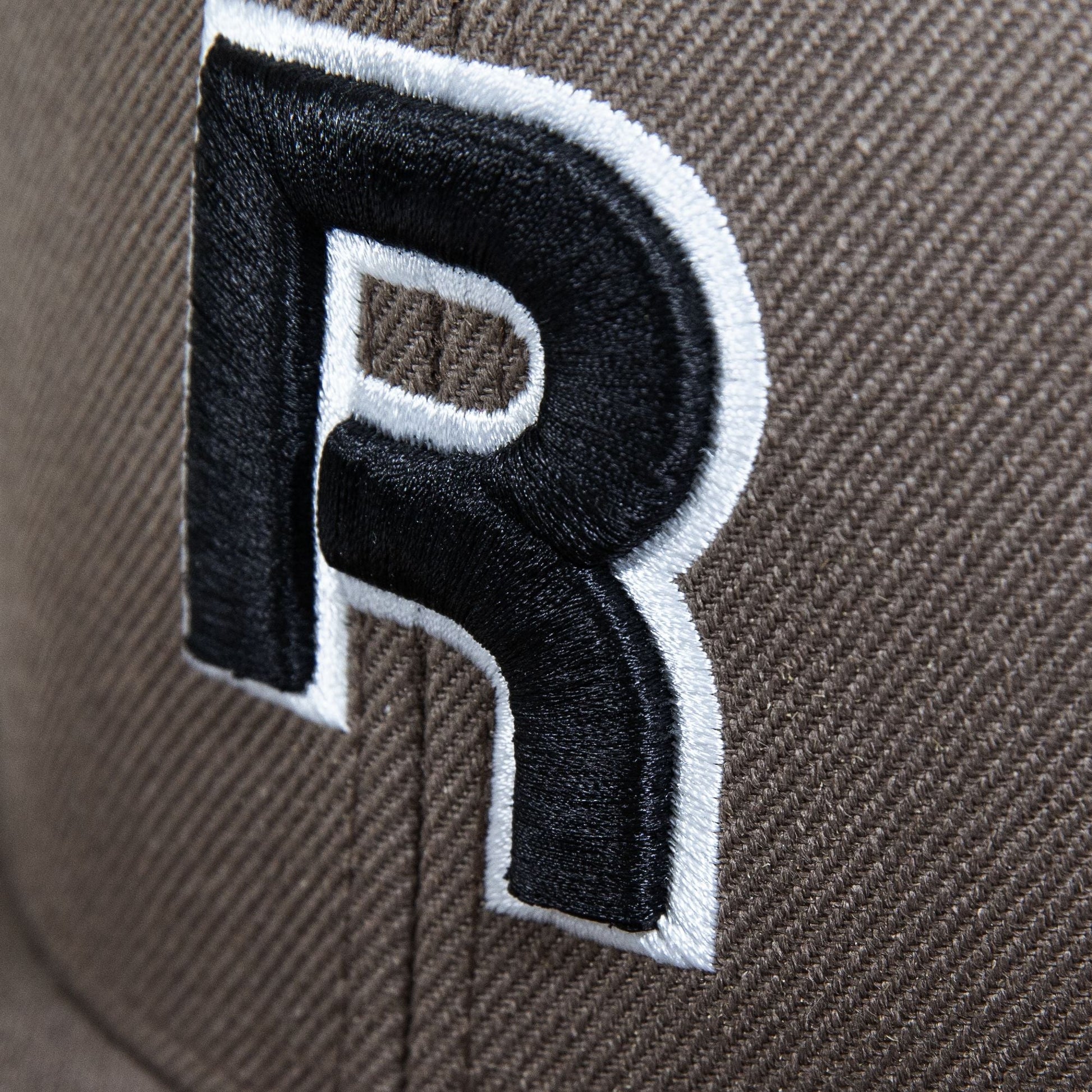 Close-up of Reebok's Identity Drop R Cap in brown, showcasing a large embroidered black "R" outlined in white, with visible stitching, texture, and a flat brim.