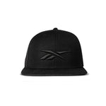 The Reebok Identity Vector Cap is a black snapback hat with a flat brim and adjustable closure, featuring an embroidered black Reebok logo on the front, shown against a white background.