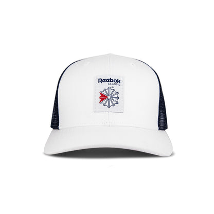 The Reebok Woven Patch Vector Trucker Cap is white with black mesh sides, a curved brim, and a navy/red logo patch. This adjustable snapback is shown on a plain white background.