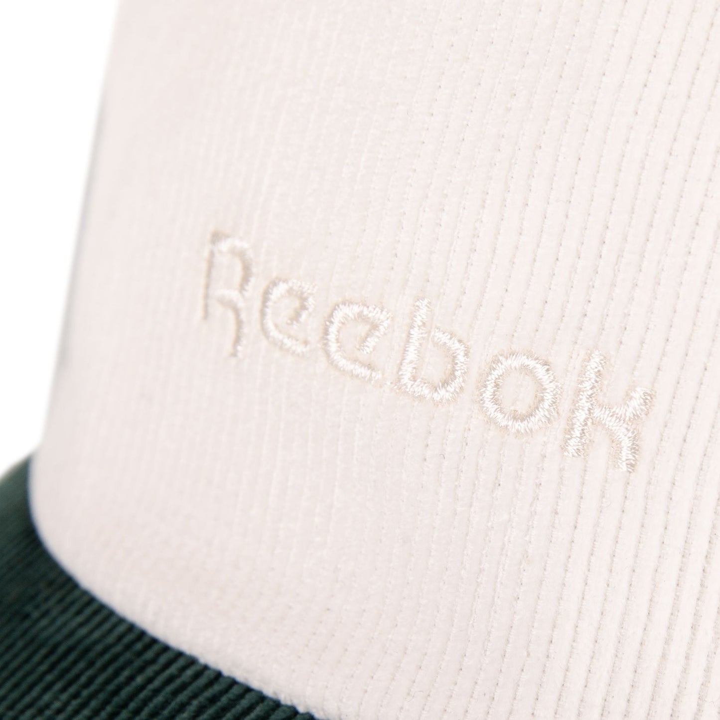 Close-up of white fabric with Reebok’s logo embroidered in white thread. The bottom edge shows a dark green or black corduroy texture, showcasing the classic style of this adjustable Reebok Logo Corduroy Cap.
