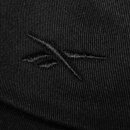 Close-up of black cotton twill fabric on the Reebok Logo S-Vector Cap, featuring a stitched abstract crossed-lines logo and textured pattern on an adjustable snapback.