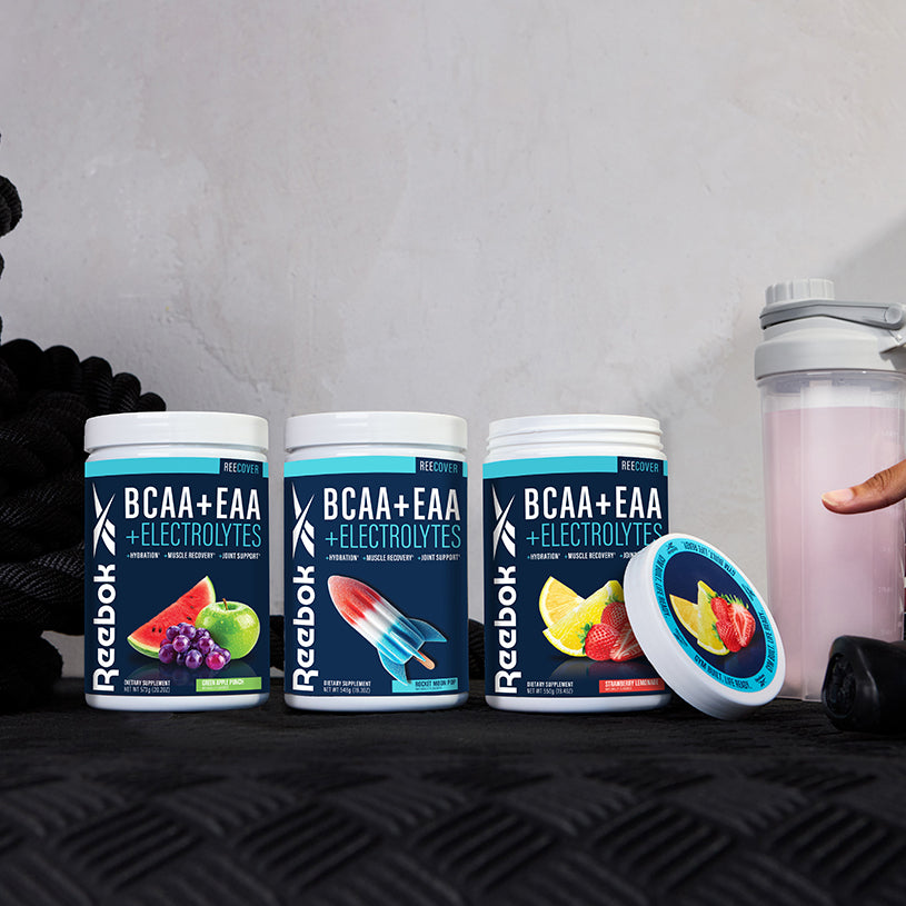 Three Reebok ReeCover BCAA + EAA + Electrolytes tubs in assorted flavors are shown on a black surface beside a shaker bottle and a hand holding its lid. These supplements support muscle recovery with essential amino acids. Ropes appear in the background.
