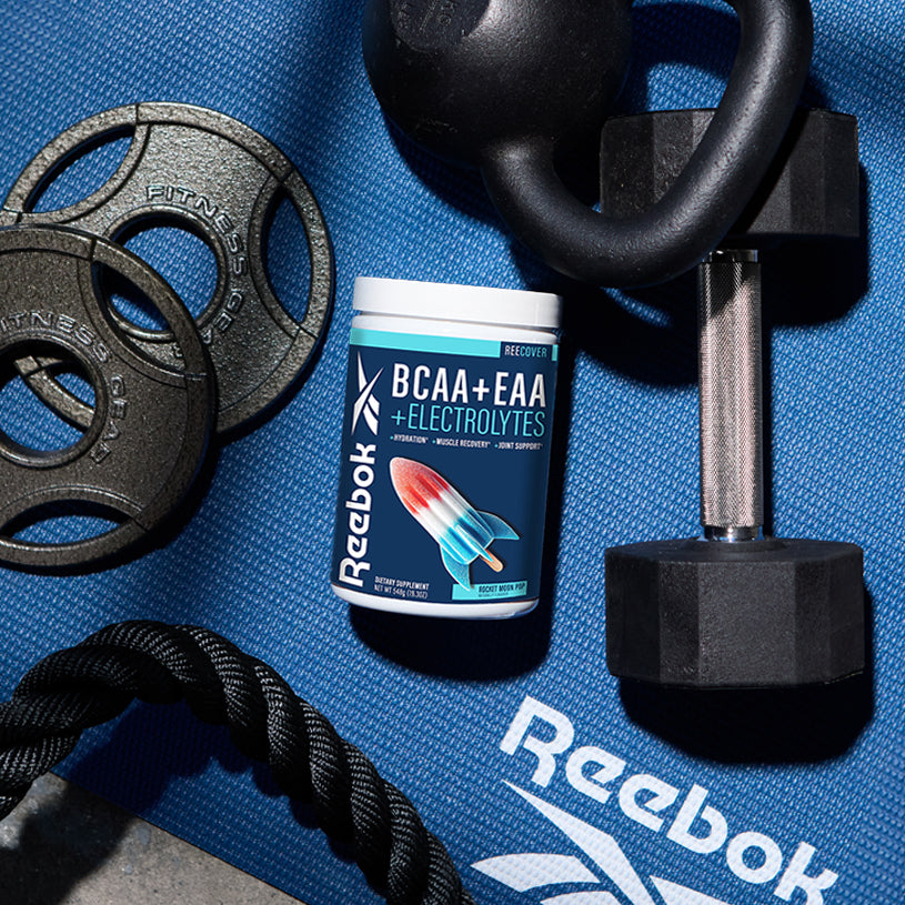 A container of Reebok ReeCover BCAA + EAA + Electrolytes, a supplement for muscle recovery, rests on a blue Reebok exercise mat next to a black dumbbell, kettlebell, gym rope, and weight plates.