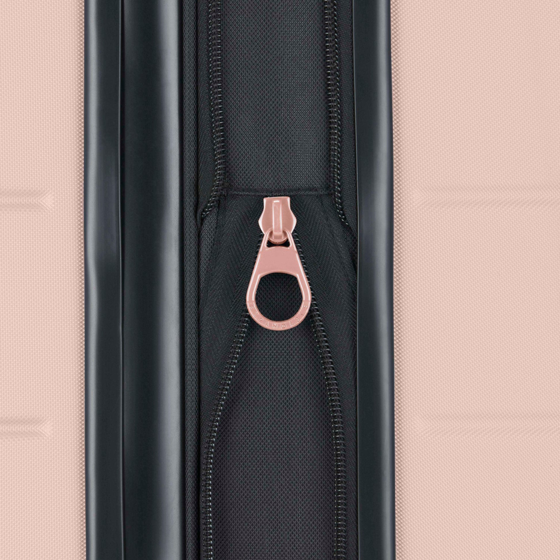 Close-up of a black zipper with a pink pull tab on a textured pink suitcase. The zipper is centered vertically between two suitcase panels.