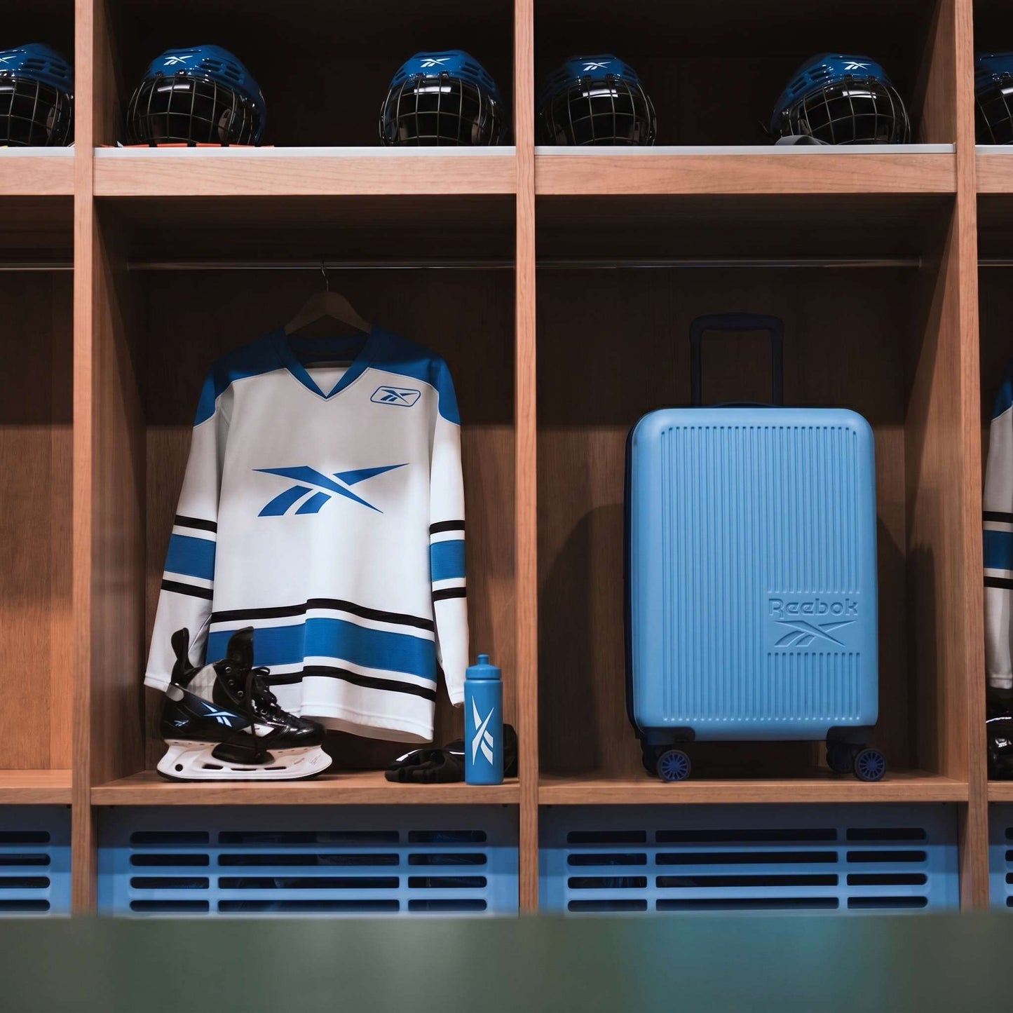 A hockey locker displays a white and blue jersey, black skates, a blue water bottle, several blue helmets on the top shelf, and the Reebok Action Duo 2 Piece Luggage Set with spinner wheels.