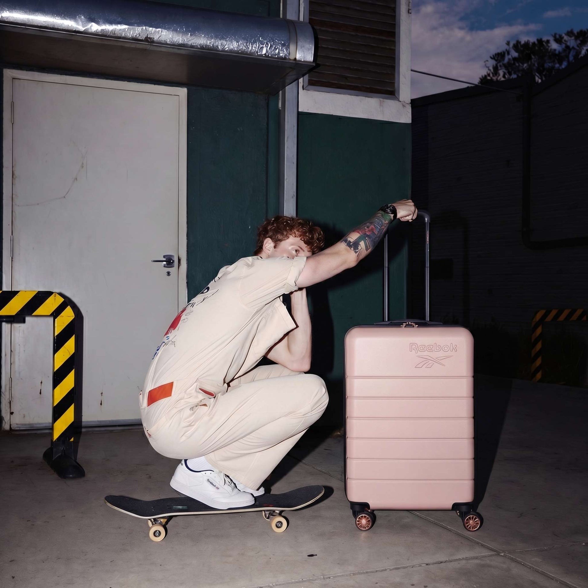 A person in a beige jumpsuit squats on a skateboard at night, holding Reebok's Captain 2 Piece Luggage Set with spinner wheels. Yellow and black safety barriers and a green wall are in the background.