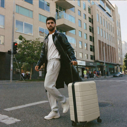 A man in a black leather coat and gray sweats crosses a city street, pulling the cream-colored Reebok Captain 2 Piece Luggage Set with spinner wheels. Modern buildings and pedestrians are visible in the background.