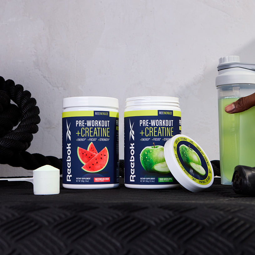 Two tubs of Reebok ReeEnergize Pre-Workout + Creatine—watermelon and green apple—sit on a gym mat with a scoop, shaker bottle, and battle rope, all set to help boost your workout endurance.