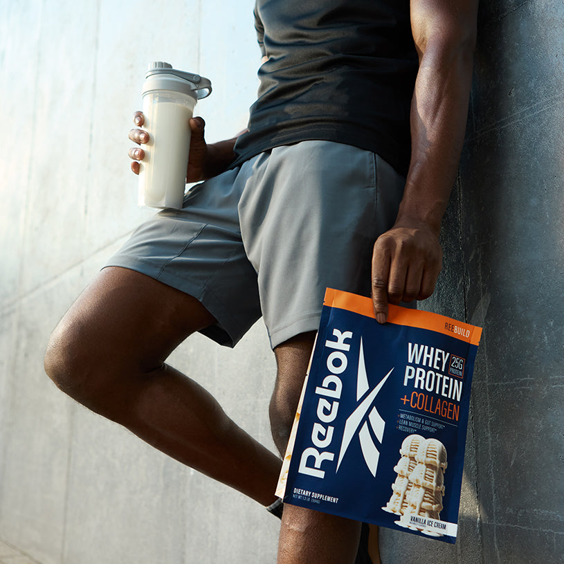 Wearing gray shorts and a black shirt, a person leans against a wall holding a shaker of milk and a Reebok ReeBuild Whey Protein + Collagen pouch, ready to support muscle recovery.
