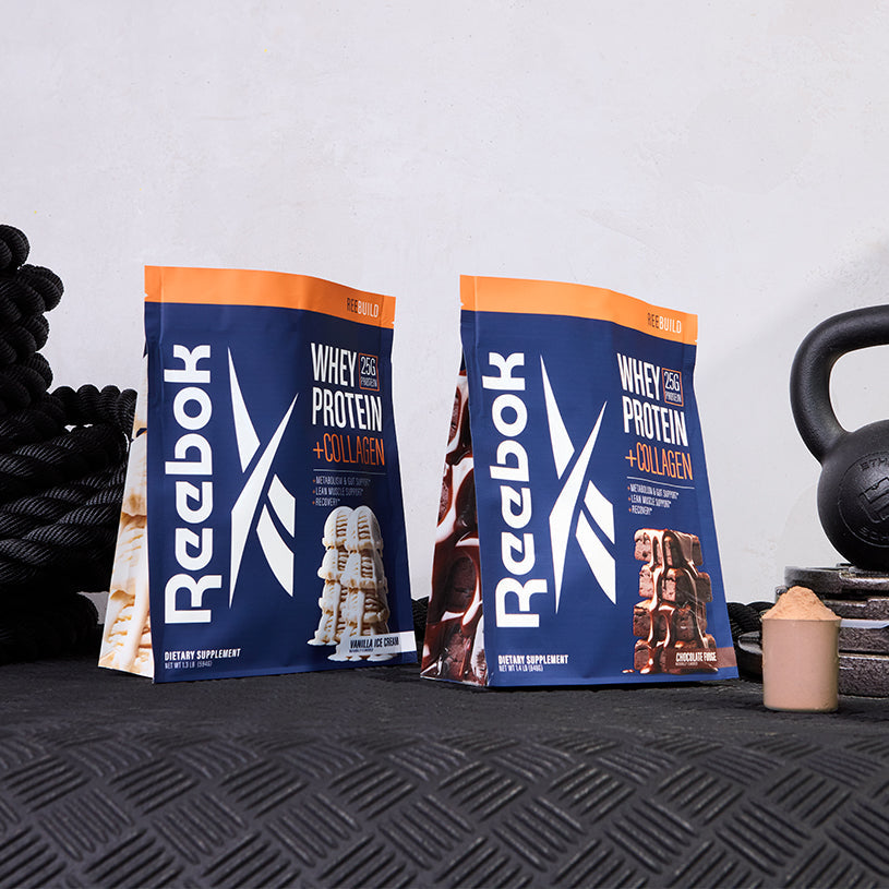 Two bags of Reebok ReeBuild Whey Protein + Collagen, one vanilla and one chocolate, rest on a textured black surface alongside a kettlebell, rope, and scoop—perfect for muscle recovery after tough workouts.