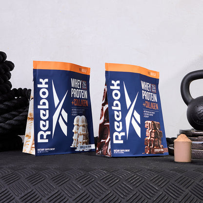 Two bags of Reebok ReeBuild Whey Protein + Collagen, one vanilla and one chocolate, rest on a textured black surface alongside a kettlebell, rope, and scoop—perfect for muscle recovery after tough workouts.