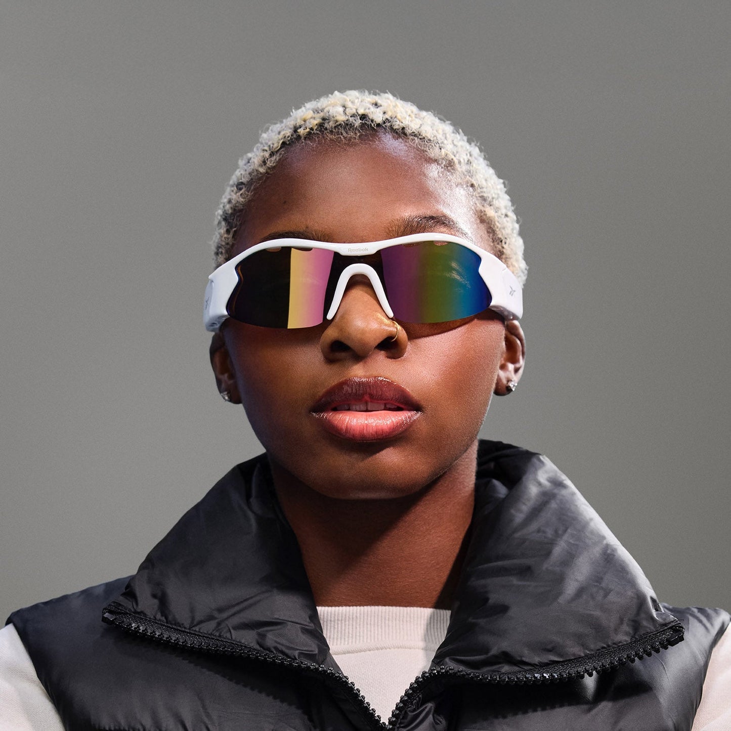 A person with short, bleached hair wears Reebok Flash Smart Audio Sunglasses and a black puffer vest over a light shirt, standing against a plain gray background.