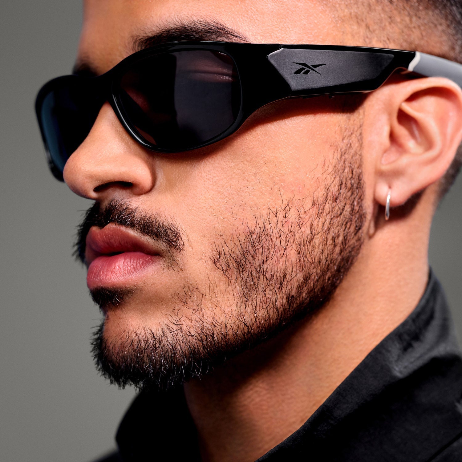 A close-up of a man with a trimmed beard and mustache, wearing Reebok Nitrous Smart Audio Sunglasses with polarized lenses and the Reebok logo visible on the side. He also wears a small hoop earring and a dark collared shirt.