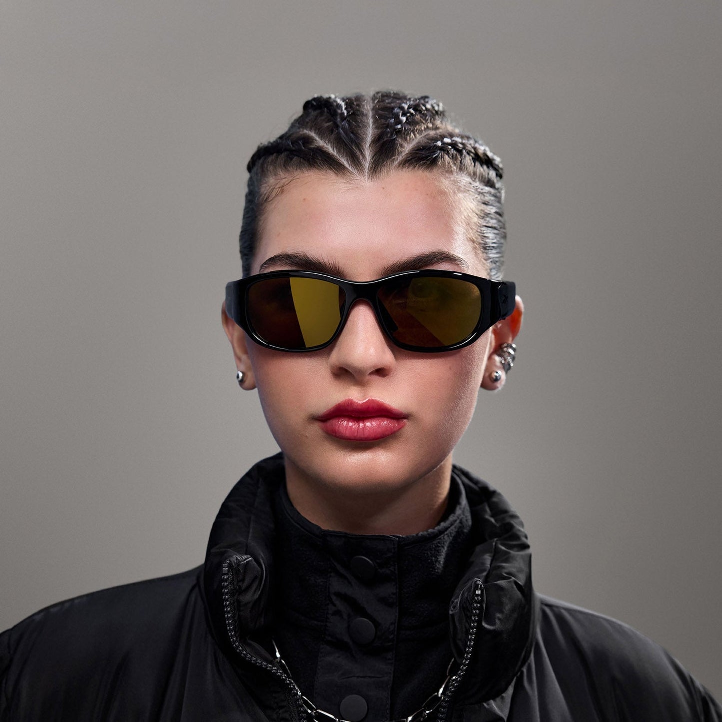 A person with braided hair wears Reebok Nitrous Smart Audio Sunglasses with polarized lenses, a black high-collar jacket, earrings, and a chain necklace while posing against a plain gray background.