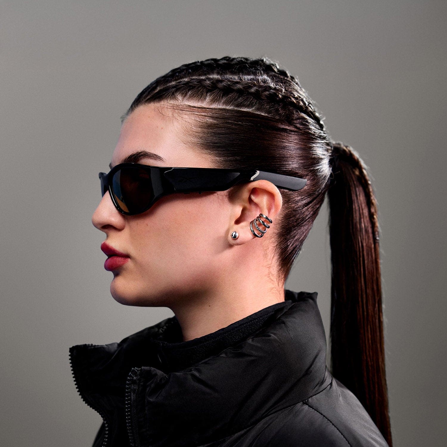 A woman in profile wears Reebok Nitrous Smart Audio Sunglasses with polarized lenses, paired with a black puffer jacket and silver jewelry. Her dark hair is in cornrow braids leading to a ponytail, set against a plain gray background.