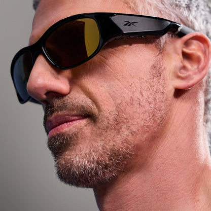Close-up of a man with short gray hair and stubble, wearing Reebok Nitrous Smart Audio Sunglasses with large tinted lenses. Only the side of his face is visible against a plain gray background.