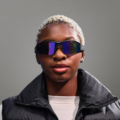 A person with short, platinum blonde hair wears Reebok Octane Smart Audio Sunglasses with polarized lenses, paired with a black puffer vest and white shirt, posing against a neutral gray background.