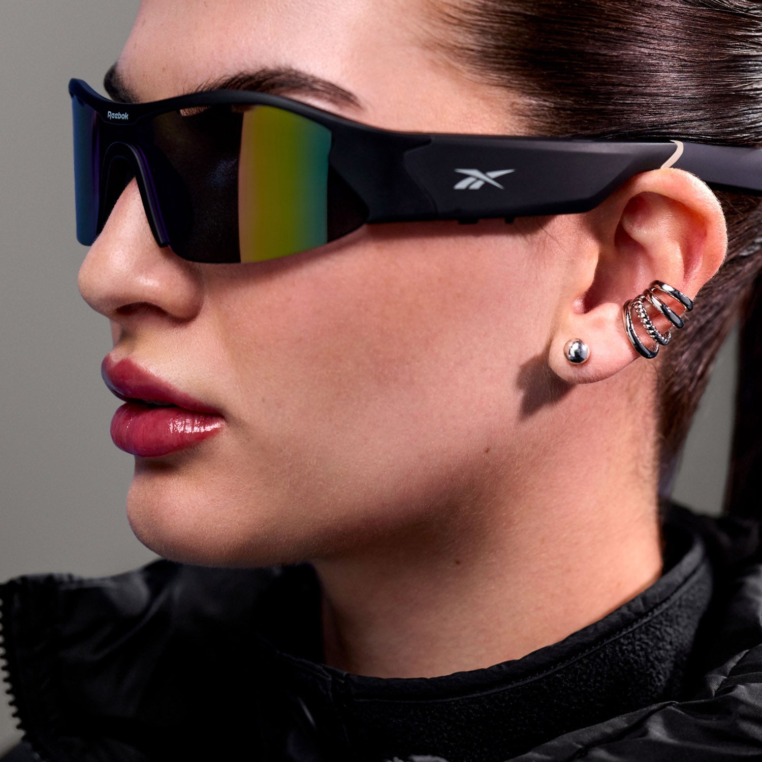 A person wearing large Reebok Octane Smart Audio Sunglasses, a black jacket, and several silver earrings—including a spiral-shaped one—is shown in close-up from a side angle.