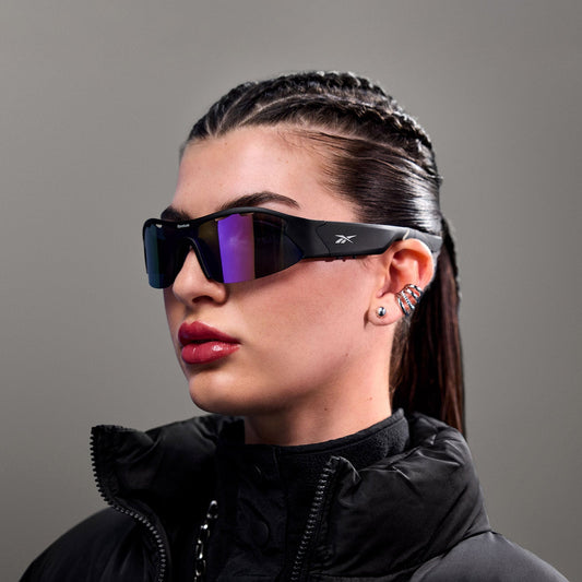A woman with braided dark hair wears Reebok Octane Smart Audio Sunglasses and a high-collared black jacket, paired with bold earrings and glossy lipstick, standing against a plain gray background.