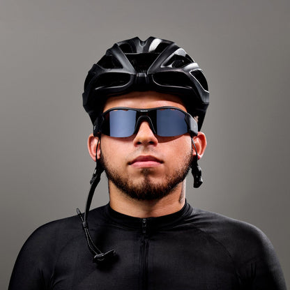 A man wearing Reebok Octane Smart Audio Sunglasses with polarized lenses, a black cycling helmet, and a black jersey stands against a gray background, looking straight ahead with a neutral expression.