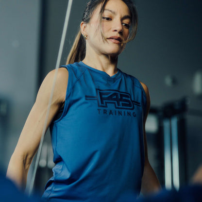 A woman wears the Women's Reebok x F45 Training Tank Top, focused as she exercises indoors. The blurred background shows gym equipment and dark tones, highlighting the Reebok branding and sweat-wicking design.