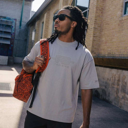 A man with long dreadlocks, sunglasses, and black shorts wears a Reebok x F45 Training Tonal Graphic T-Shirt and carries a red patterned backpack over his shoulder while walking outside near a brick building.