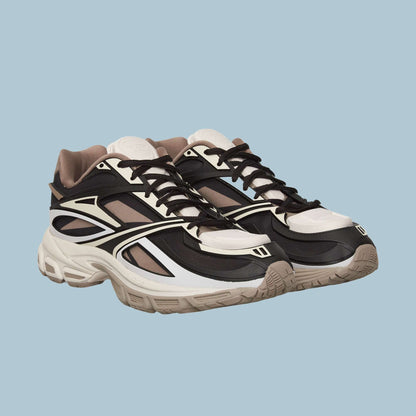 The Reebok Premier Road Modern Shoes feature black, white, and beige panels, thick soles, and black laces for a futuristic look, shown on a light blue background.
