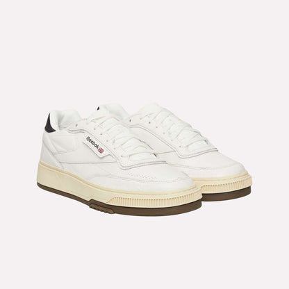 A pair of white Reebok sneakers with black detailing on the heel, off-white midsoles, gum rubber outsoles, and the Reebok logo on the side. The shoes have a retro design with lace-up fronts.
