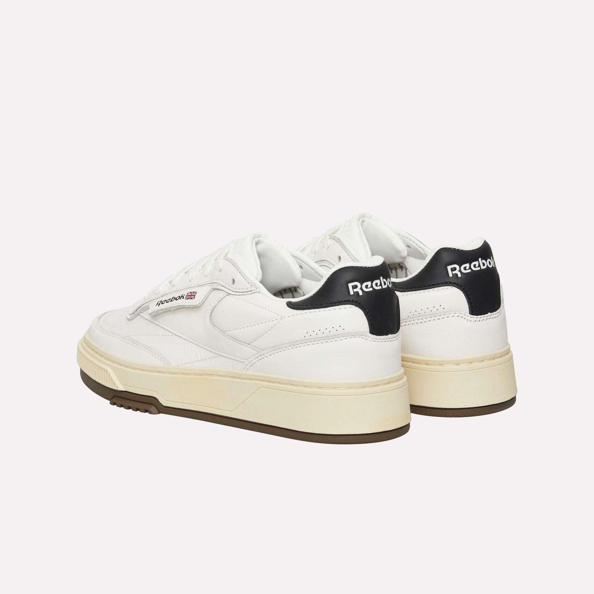 A pair of white Reebok sneakers with black heel tabs, gum-colored outsoles, and the Reebok logo visible on the side and back. The shoes are positioned at an angle, showing both the side and back.