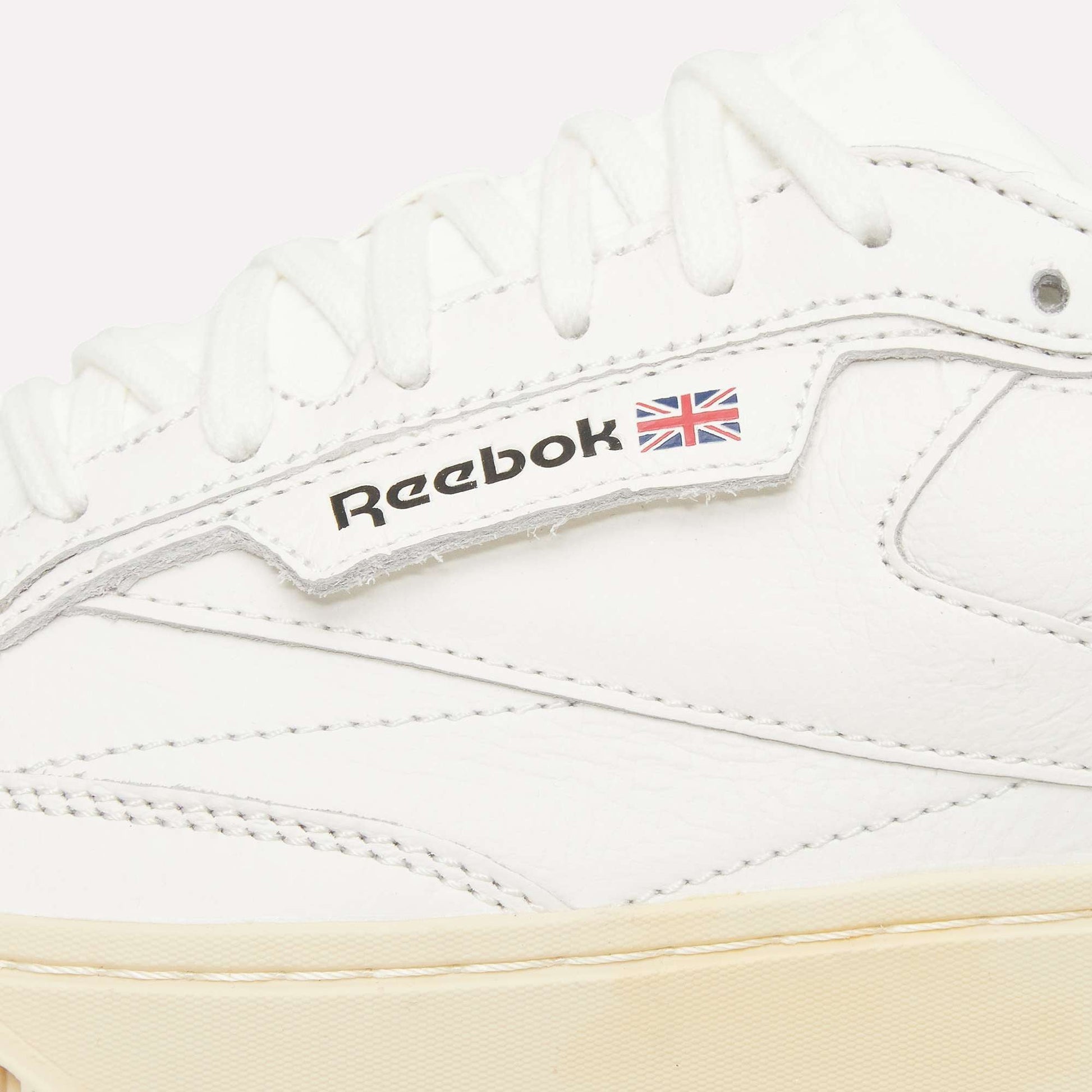 Close-up of a white Reebok sneaker, showing white laces, stitching, and the Reebok logo with a small Union Jack flag on the side. The sole is a light beige color.