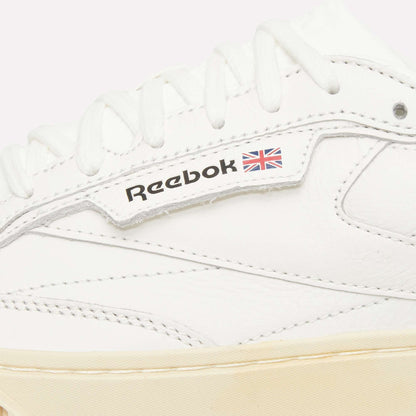 Close-up of a white Reebok sneaker, showing white laces, stitching, and the Reebok logo with a small Union Jack flag on the side. The sole is a light beige color.