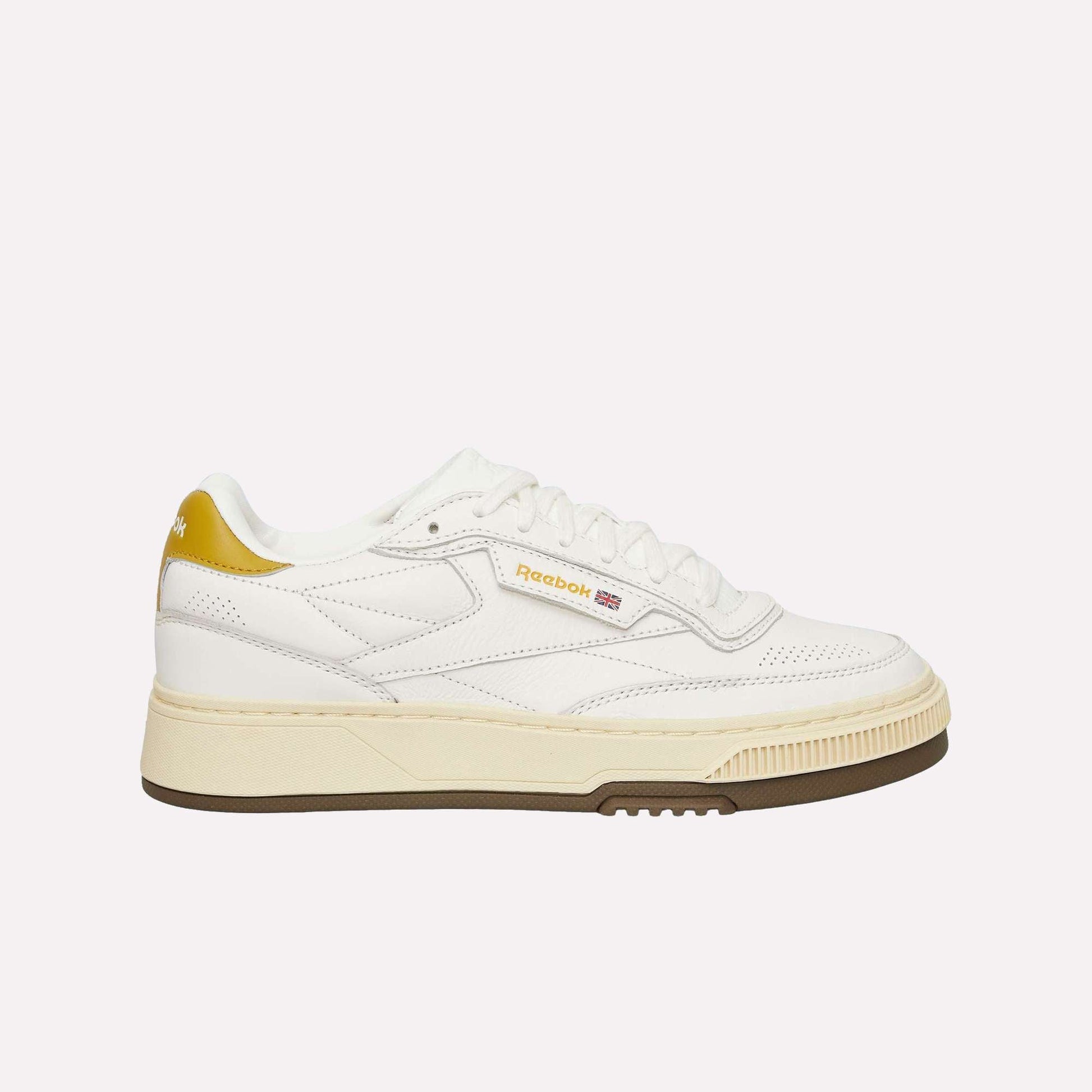The Reebok Club C LTD Shoes feature a white upper, gold heel and logo accents, a beige midsole, and a brown outsole. Shown on a light blue background, these classic tennis sneakers offer timeless style.