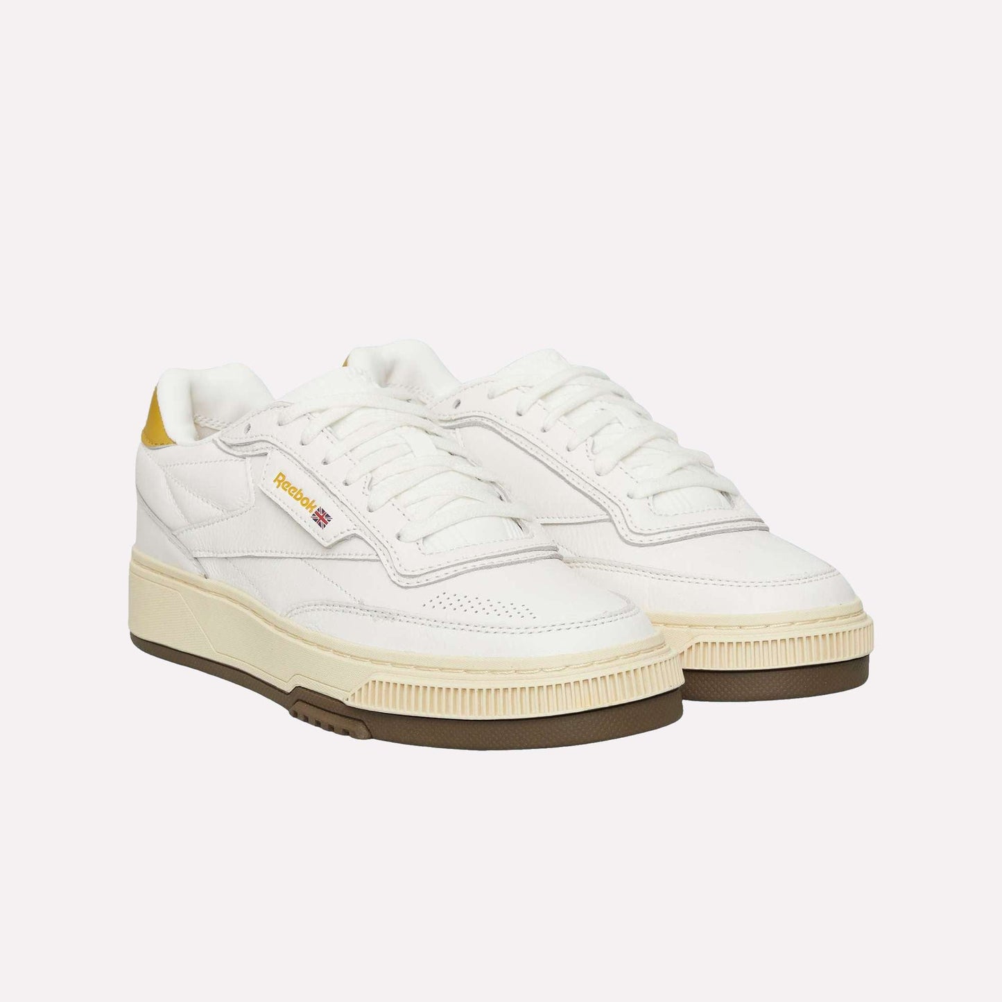 The Reebok Club C LTD Shoes feature a white upper with yellow heel accents, a beige midsole, gum outsole, and the signature Reebok logo on the side—lifestyle classics shown against a light blue background.