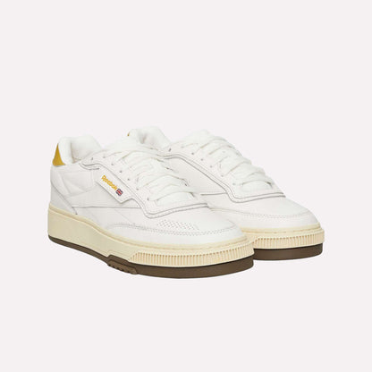 The Reebok Club C LTD Shoes feature a white upper with yellow heel accents, a beige midsole, gum outsole, and the signature Reebok logo on the side—lifestyle classics shown against a light blue background.