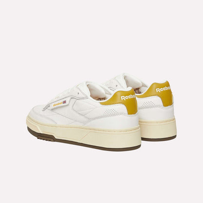 A pair of Reebok Club C LTD Shoes in white with yellow heel accents and beige soles are shown against a light blue background, angled to display the side and back of this classic lifestyle tennis shoe.