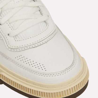 Close-up of the Reebok Club C LTD Shoes—a white leather tennis shoe with perforated details, beige rubber sole, white laces, and textured stitching—set against a light blue background.