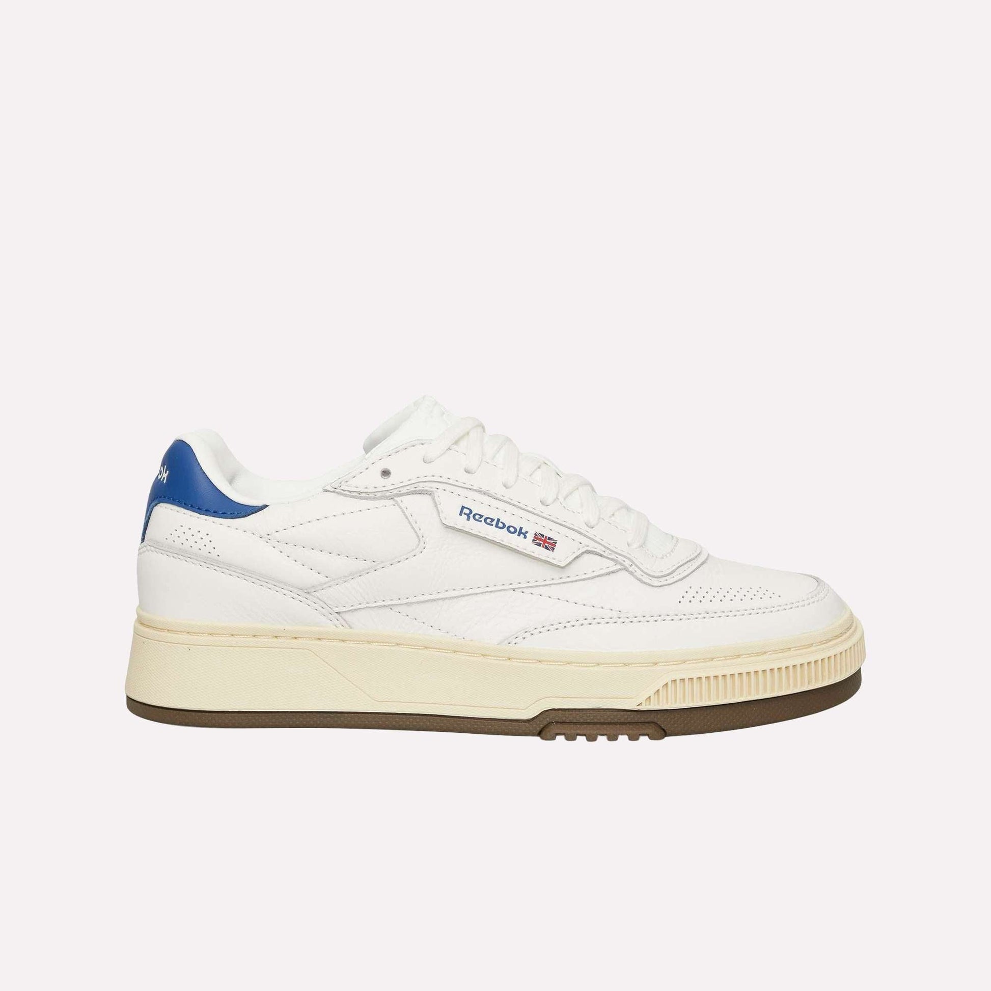 The Reebok Club C LTD Shoes are classic white tennis shoes with a blue heel detail, beige midsole, brown outsole, and feature the Reebok logo plus a small Union Jack flag on the side.