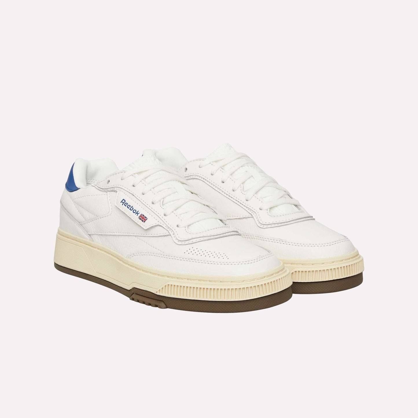 A pair of Reebok Club C LTD Shoes in white with blue heel accents, a beige midsole, and brown outsole is displayed on a white background. The Reebok logo and British flag detail complete this classic lifestyle tennis shoe.