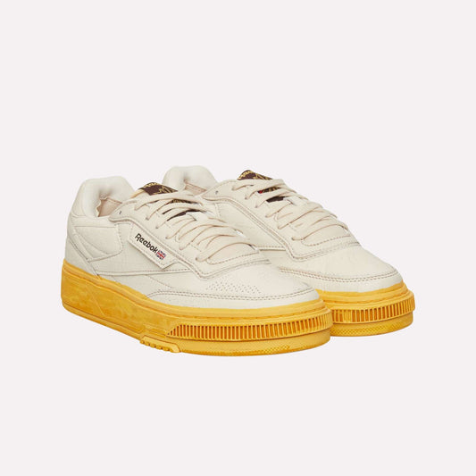 The Reebok Club C LTD Shoes are cream-colored with yellow soles, white laces, and a retro-inspired design. They feature the Reebok logo and Union Jack on the side, blending classic tennis shoe style with modern flair.