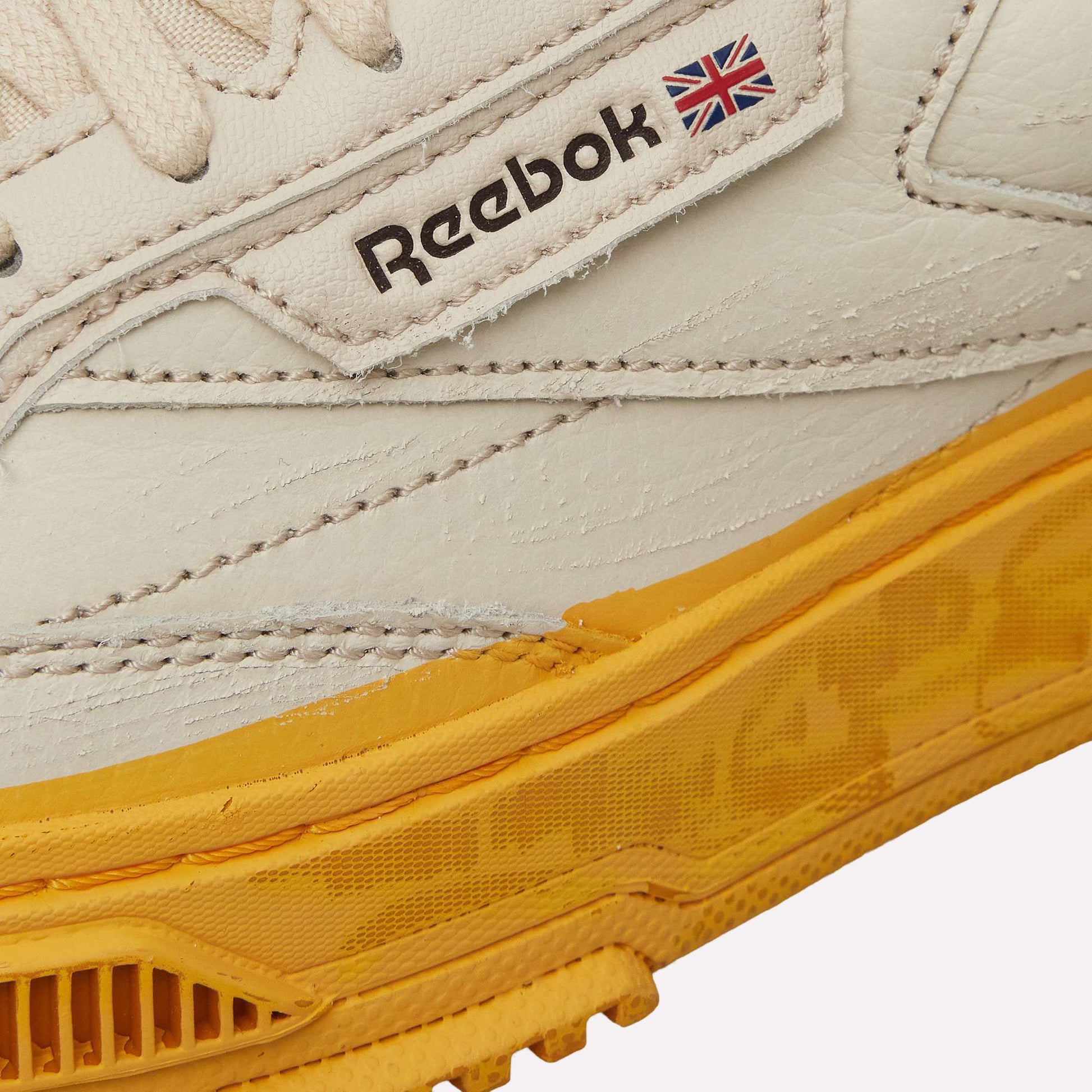 Close-up of the Reebok Club C LTD Shoes in white, showcasing the Reebok logo, UK flag, and a textured yellow rubber sole. Classic stitching details accent the leather upper for this timeless lifestyle sneaker.