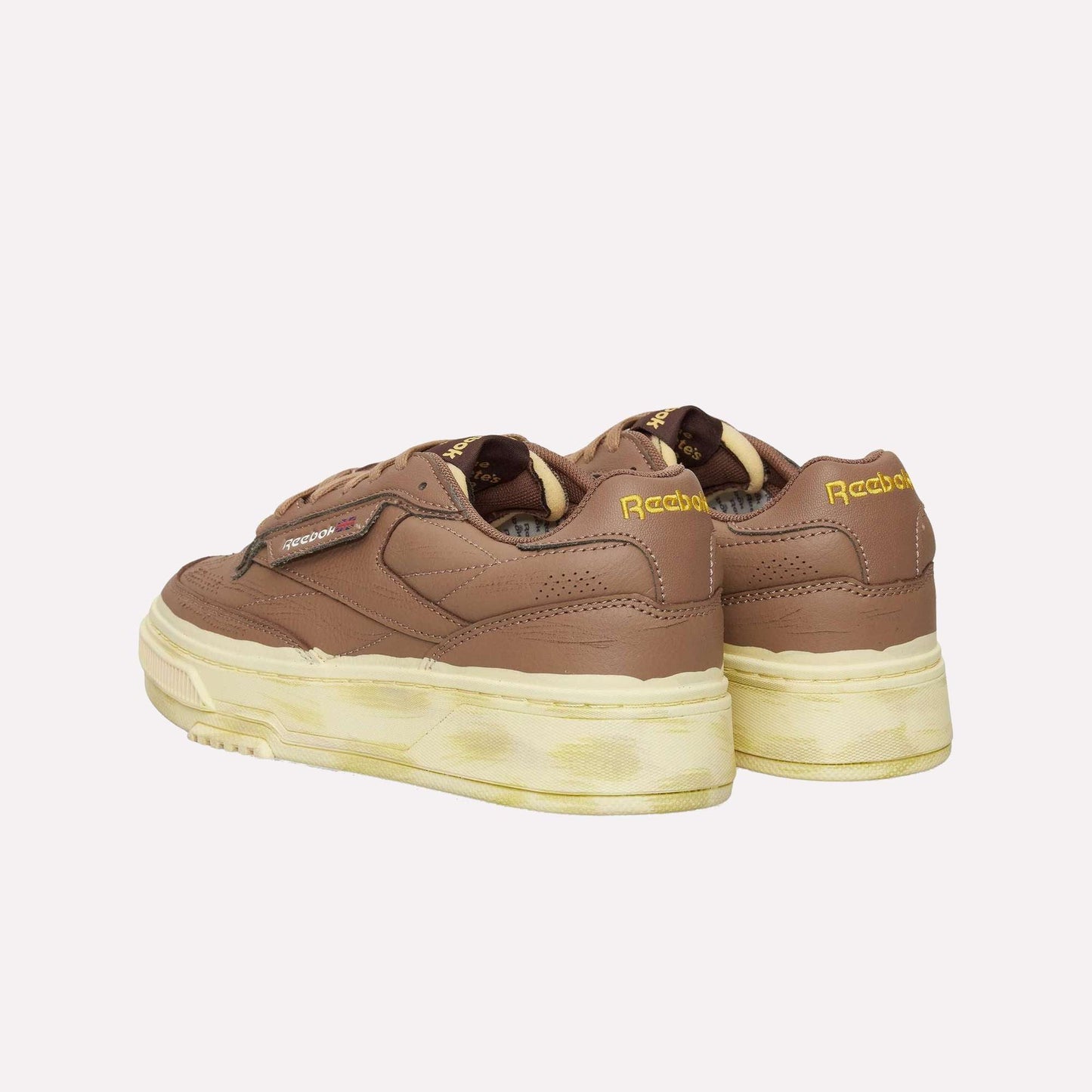 A pair of Reebok Club C LTD Shoes in brown with yellow logo accents and thick off-white soles, featuring a vintage-inspired, slightly worn look—shown from both back and side angles.